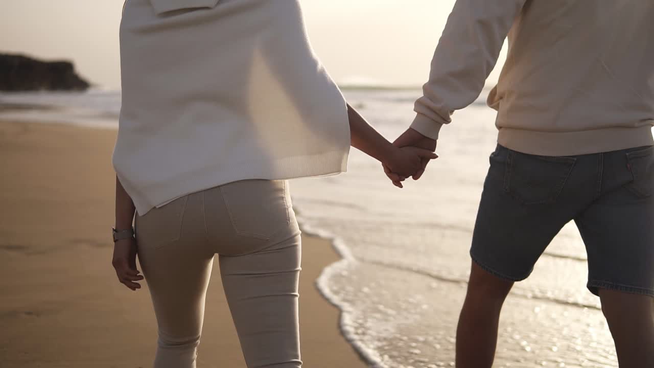 Backside view of young couple in love holding hands walking on the beach in casualclothes. Background sunset in sea. Slow Motion