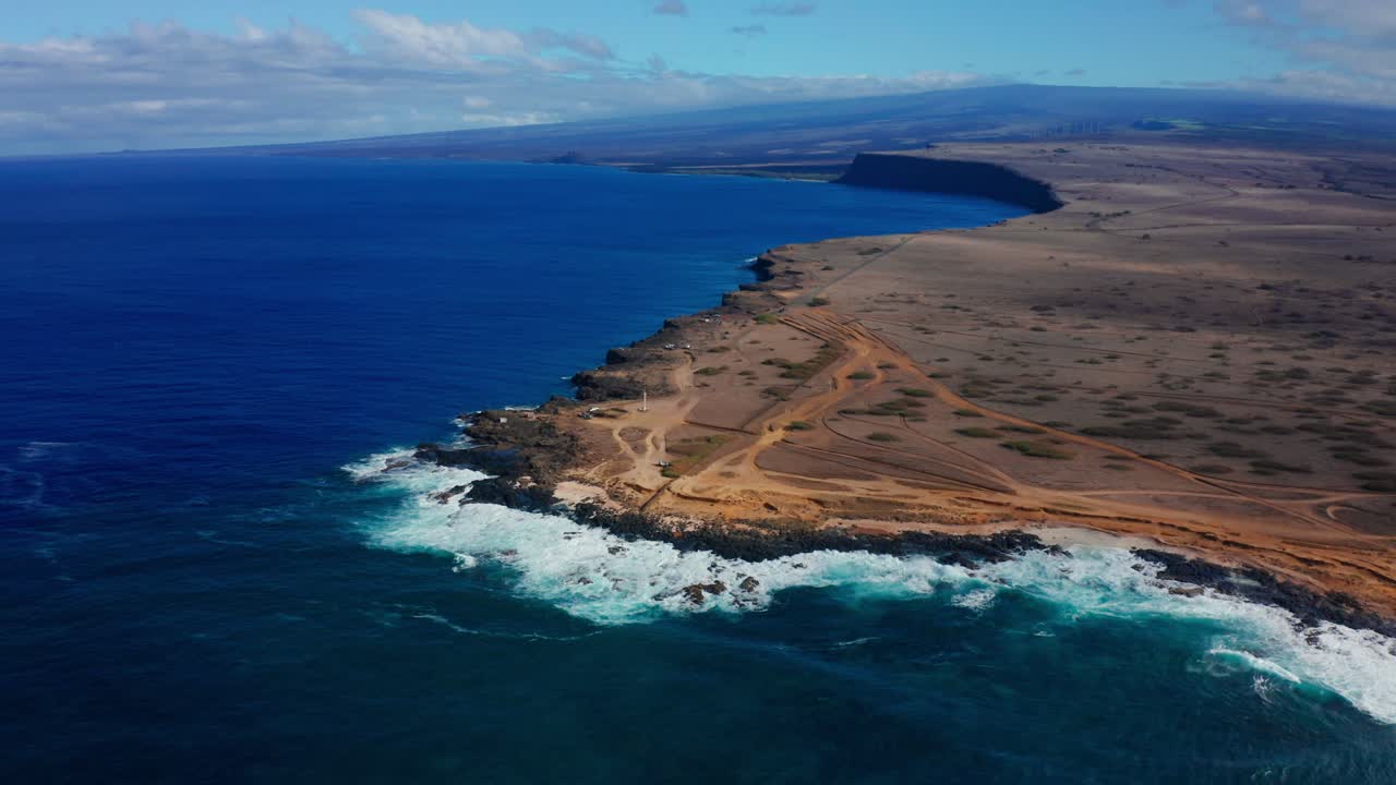 Foaming waves pound a rocky desert shoreline that stretches into the distance, framed by vivid blue ocean and a bright expansive sky for a wild coastal scene.