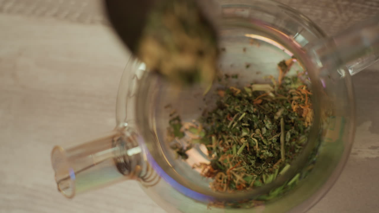 Ervas secas em infusão numa chaleira de vidro, vista superior aproximada de camomila e hortelã, herbalista mexendo com uma colher, luz quente de cozinha, ambiente relaxante de spa, vapor suave subindo da mistura.