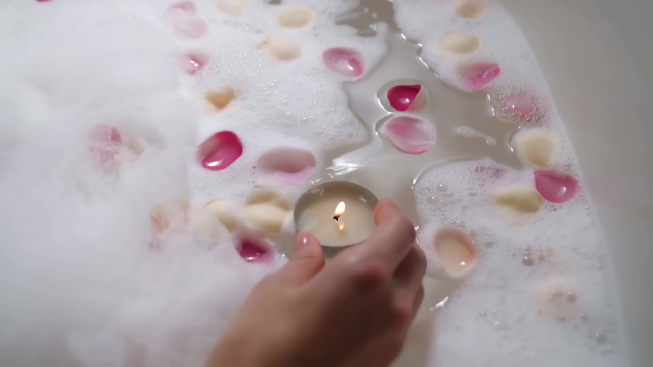 Close-up of a hand holding a small lit candle near the surface of a white foamy bath, with scattered pink and cream rose petals creating a calm spa atmosphere