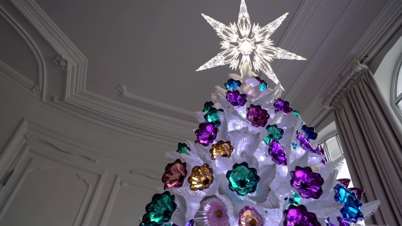 Elegant white Christmas tree adorned with vibrant floral ornaments and luminous star, positioned in spacious, refined room featuring expansive windows and pristine walls