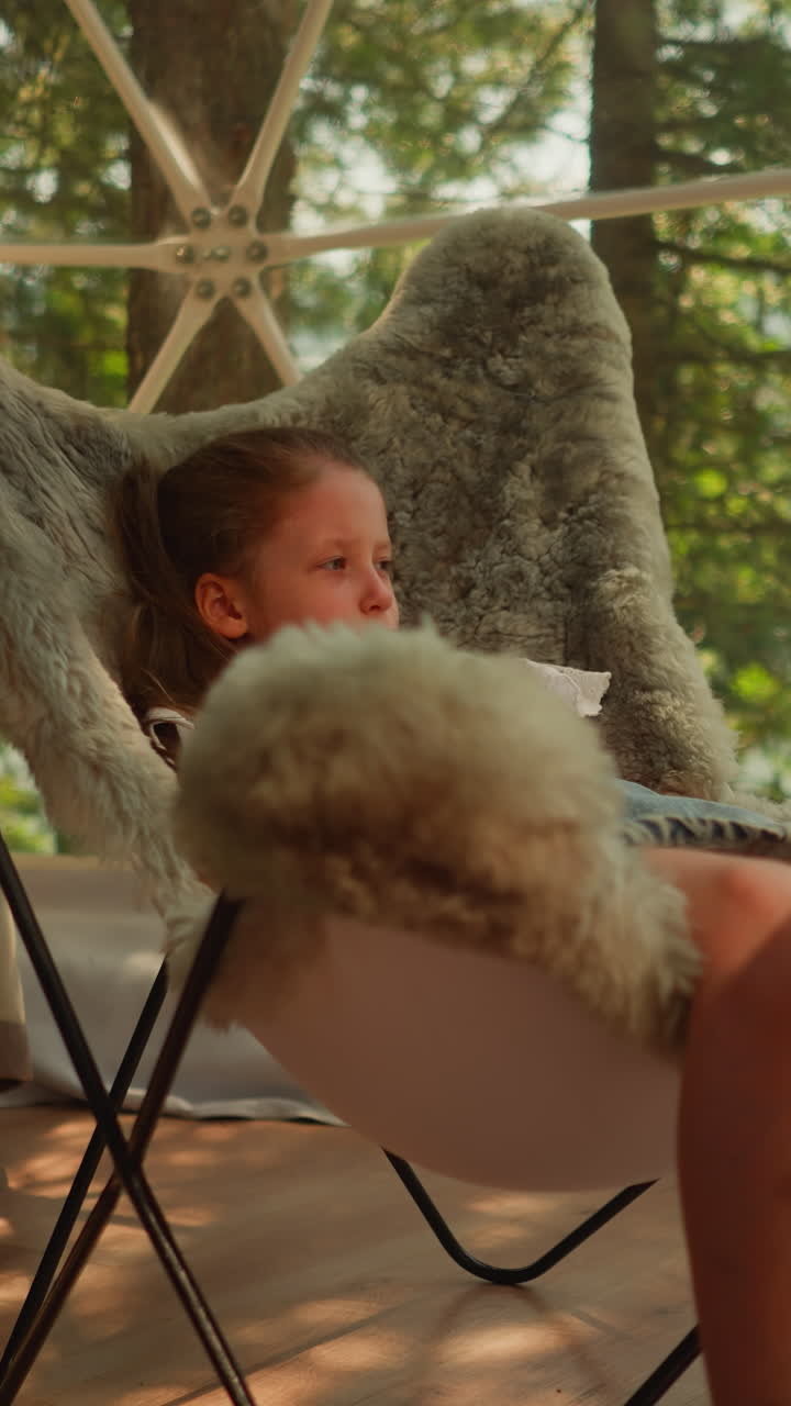 niña pensativa observa a los pájaros fuera de la ventana balanceando las piernas mientras está sentada en una silla de piel. niño estudia la naturaleza observando desde el interior. alojamiento en glamping