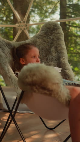 pensive little girl watches birds outside window swinging legs while sitting in fur chair. kid studies nature watching from indoors. accommodation in glamping