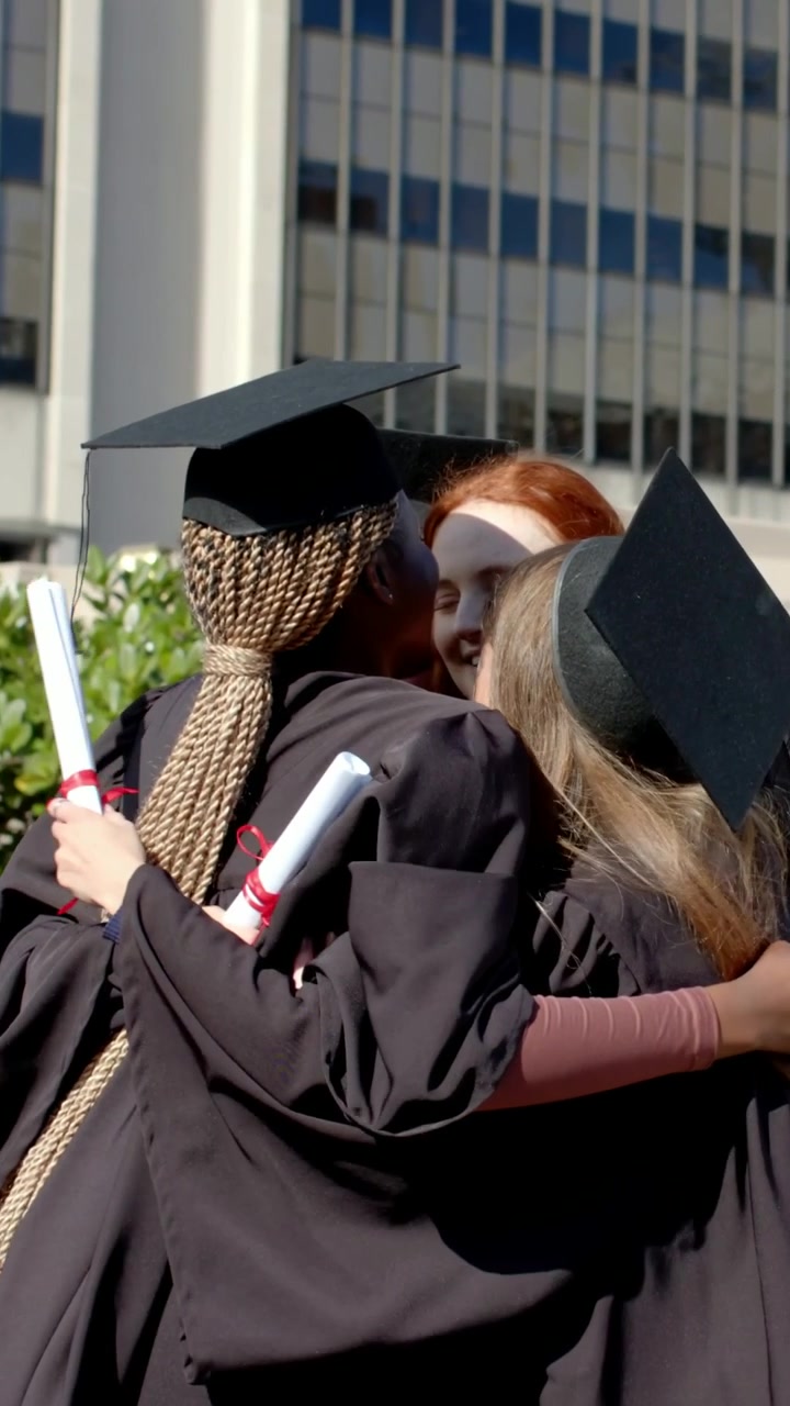 Video verticale: Quattro laureati con trecce si abbracciano, indossando le toghe dopo la cerimonia in piazza e tenendo in mano i diplomi.