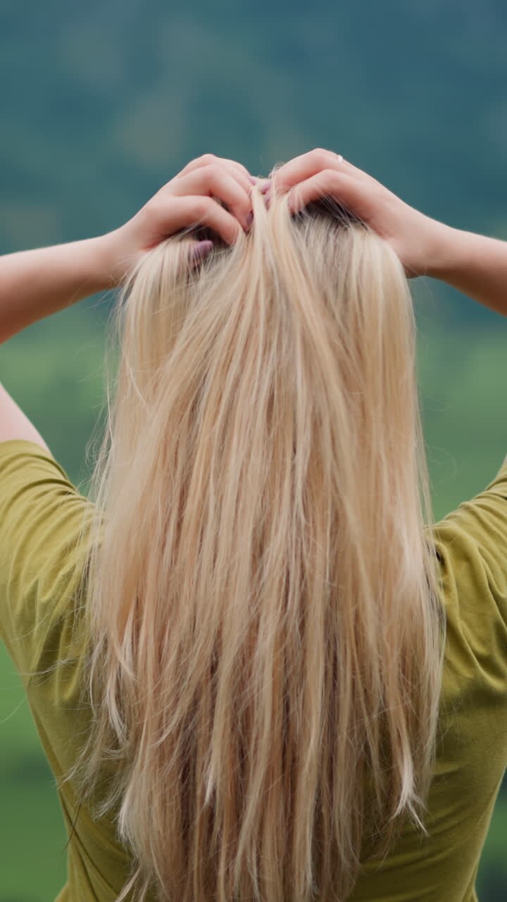 Lady traveler adjusts long hair by hands standing over green valley surrounded by forestry mountains on overcast day close view slow motion