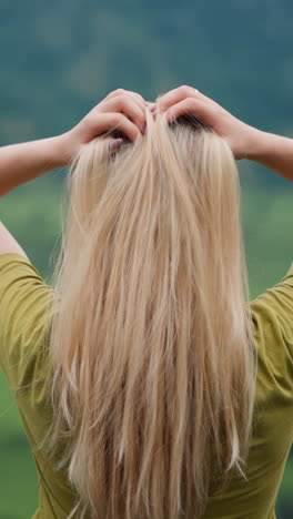 lady traveler adjusts long hair by hands standing over green valley surrounded by forestry mountains on overcast day close view slow motion