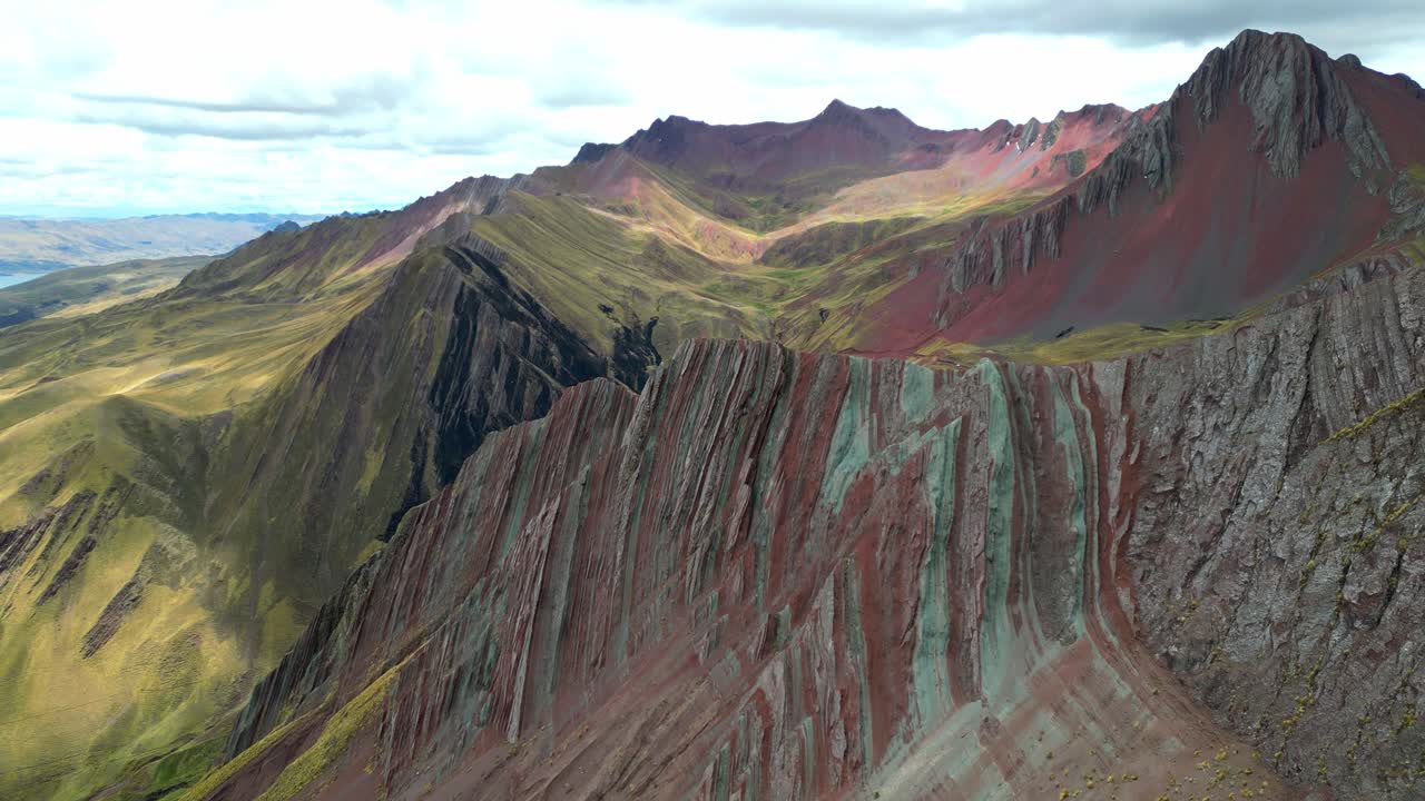 Filmato aereo che sorvola le vivaci creste minerali di Palccoyo. Stratificazioni colorate increspano le cime, sullo sfondo delle Ande innevate in lontananza.