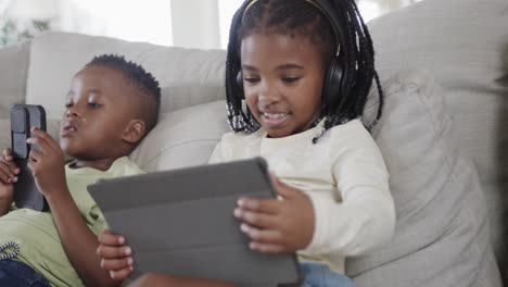 happy african american brother and sister using tablet and smartphone on sofa at home, slow motion