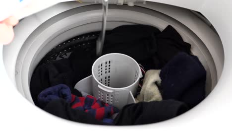 hand reaching opening cap on white washing machine appliance at home and pouring liquid into drum with full load, colorful socks and dirty wash