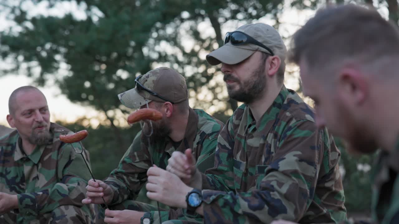 hombres militares en uniformes moro relajándose después de un turno de la tarde en la base en el campo, sentados en cajas afuera y preparando una fogata, recogiendo salchichas en palos, preparando el almuerzo, hablando, riendo