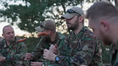 military men in moro uniforms relaxing after an afternoon shift at base in the field, sitting on crates outside and preparing campfire, scooping sausage on sticks, preparing lunch, talking, laughing