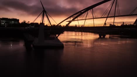 Static-drone-shot-of-South-Bank's-Goodwill-Bridge-at-sunrise,-with-bridge-silhouetted-over-orange-sky