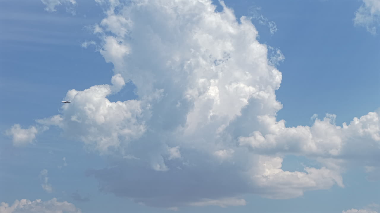 Static shot frames distant passenger plane passing before textured cumulus cloud against a clear blue sky