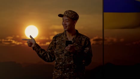 soldado con teléfono inteligente al atardecer, bandera de ucrania en el fondo