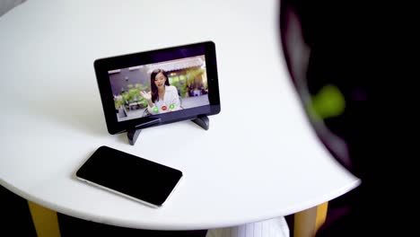 vista trasera de una joven musulmana sentada en una mesa en la habitación negociando usando una tableta y una aplicación de video llamada, agitando la mano haciendo un gesto de saludo, teniendo una reunión en línea de video llamada con una amiga