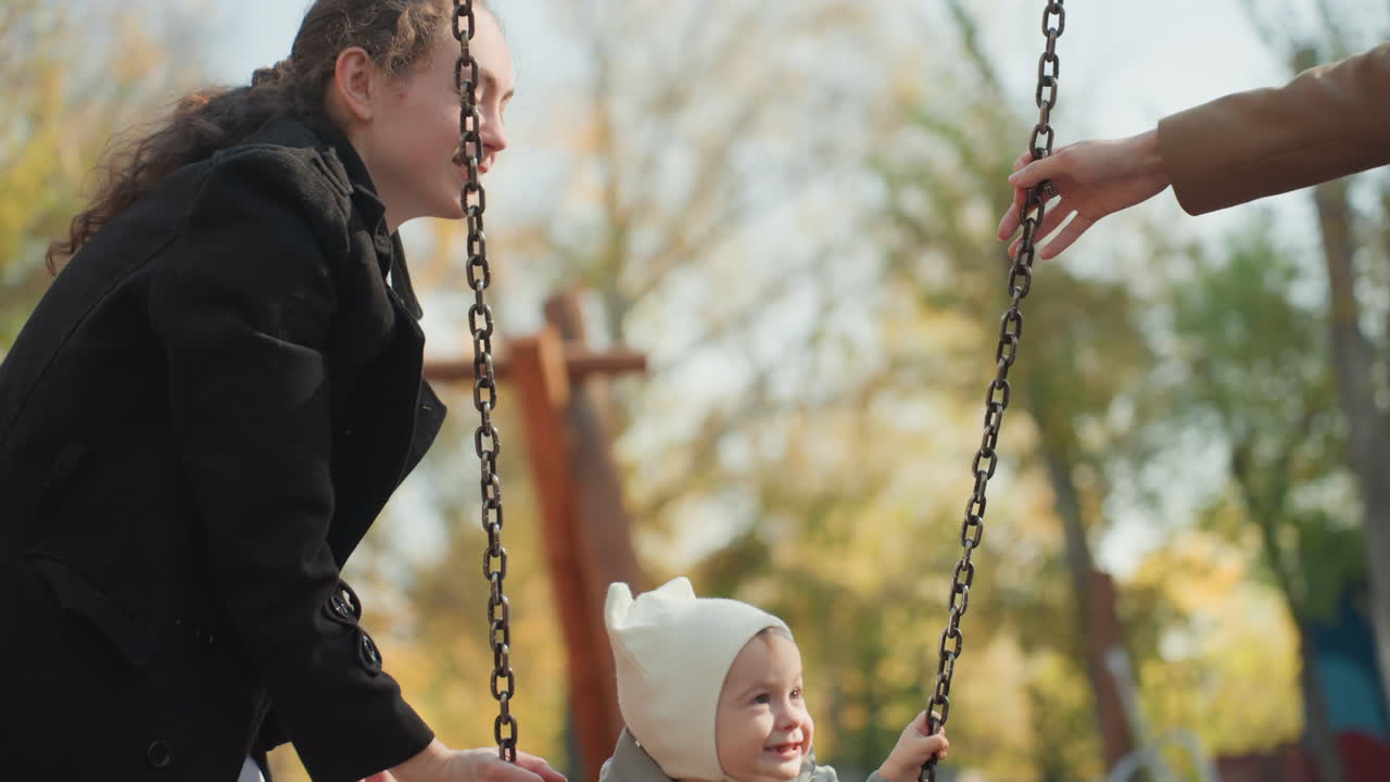 Madre di etnia mista spinge l'altalena del bambino, parco autunnale, interazione genitoriale sorridente, scena familiare coniugale intima che mostra cura delicata, spinta giocosa e contatto visivo, ruolo del genitore come custode e insegnante, calda.