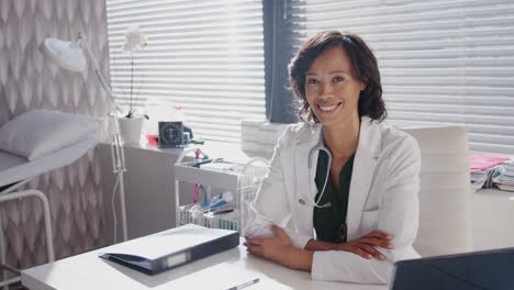 portrait of smiling female doctor wearing white coat with stethoscope sitting behind desk in office