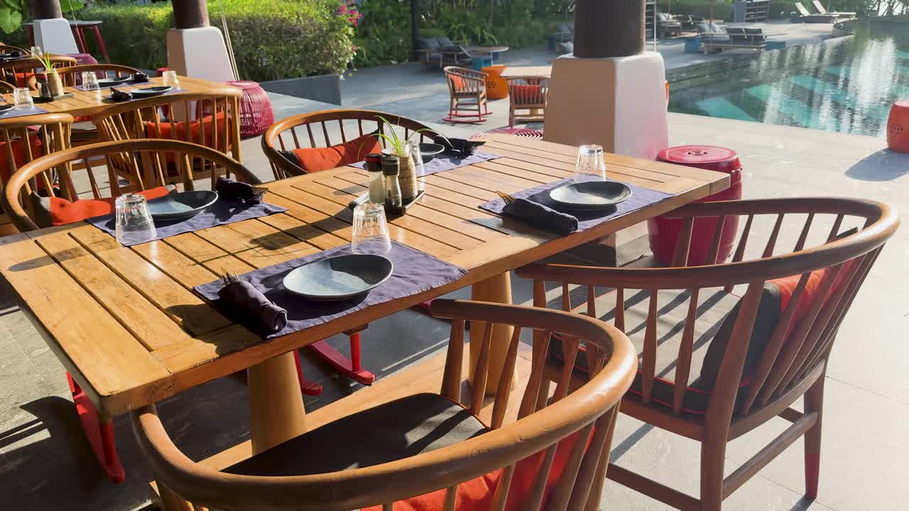 Empty outdoor dining area by a pool in Phuket, Thailand. Bright sunlight, wooden furniture, serene atmosphere captured in 16 seconds