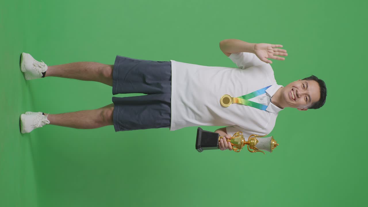 황금 메달과 트로피를 들고 있는 아시아 남성의 몸이 가득 차서 손을 흔들고 카메라를 향해 미소 짓으며 스튜디오의 녹색 스크린 배경에서 첫 번째 우승자로