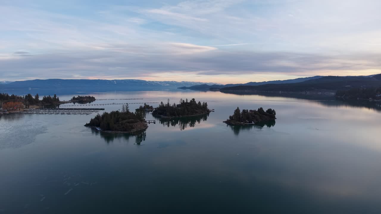 Una serena vista aérea del lago Flathead cerca de Whitefish, Montana, muestra aguas tranquilas salpicadas de pequeñas islas boscosas, montañas distantes y un cielo en tonos pastel