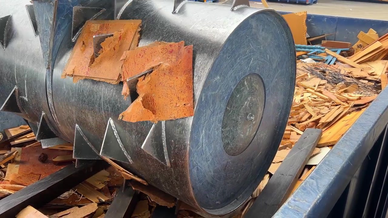 Tamburo compattatore di rifiuti che schiaccia montanti di legno in un contenitore per il riciclo presso un centro di raccolta rifiuti.