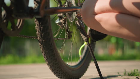 low-angle view of individual squatting beside her bicycle, intently pumping air into the tire with green nozzle on sunny day, bicycle in sharp focus, background softly blurred with fence and trees