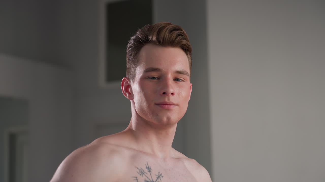 shirtless young man with tattoo on chest proudly displaying upper body muscles while smiling and gazing confidently toward camera in bright indoor setting with smooth background