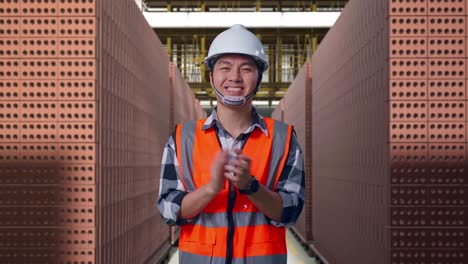 asian male engineer with safety helmet smiling and clapping his hands while standing with red brick packed in stacks are stored