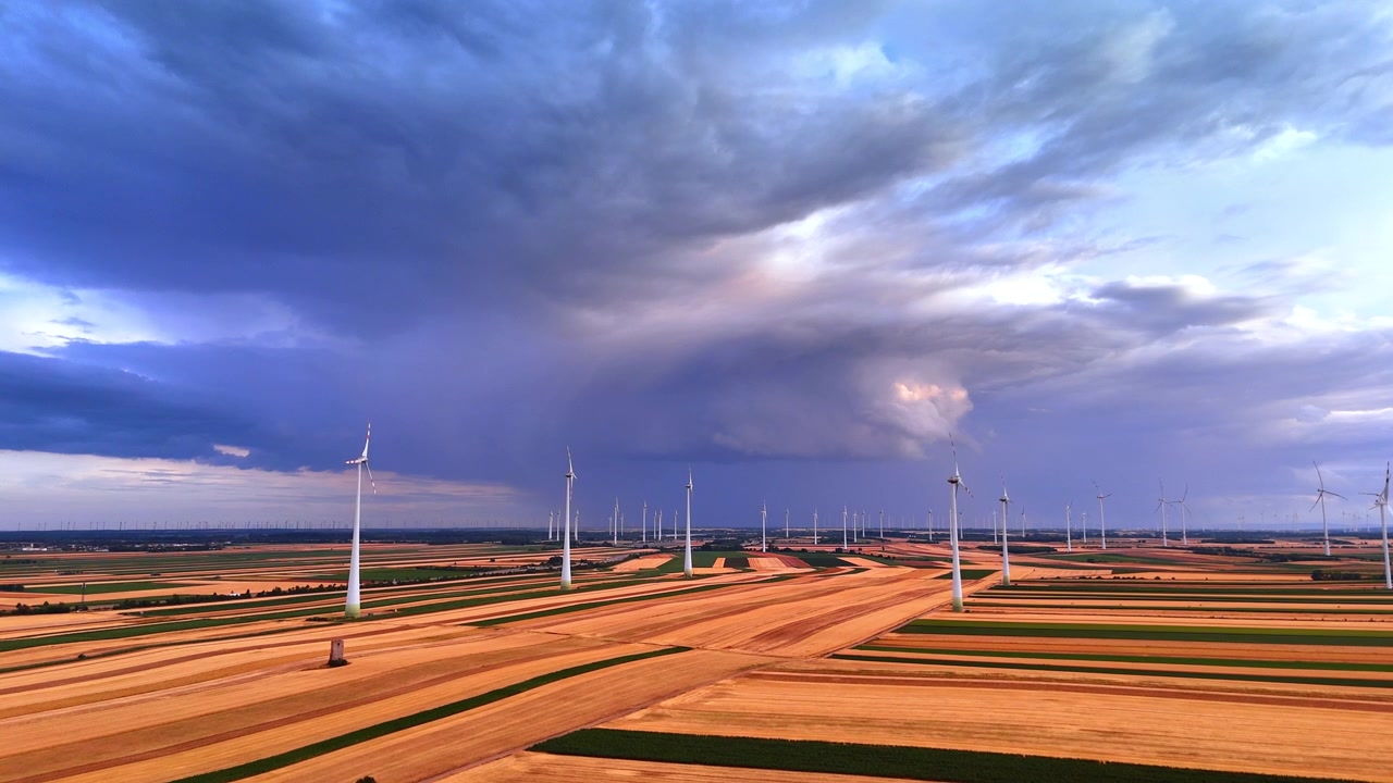 Molteplici turbine eoliche in funzione attraverso campi agricoli a strisce. Ampia prospettiva aerea di un parco eolico sotto drammatiche nuvole temporalesche blu scuro e un cielo coperto.