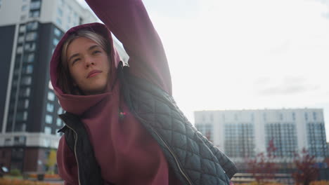 young woman in hoodie and quilted jacket stretches side muscles outdoor under bright sun against backdrop of tall city buildings and autumn leaves on paved plaza during energetic workout session