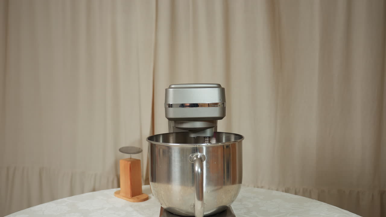 Electric mixer actively blending ingredients in stainless steel bowl placed on round table covered with patterned cloth, accompanied by small wooden kitchen tool, with beige curtain background