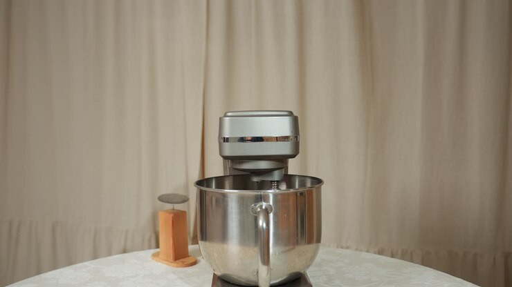 Electric mixer actively blending ingredients in stainless steel bowl placed on round table covered with patterned cloth, accompanied by small wooden kitchen tool, with beige curtain background