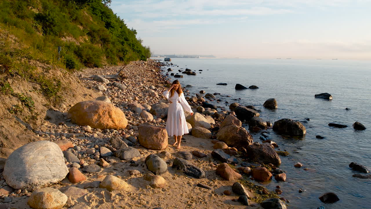 ⁇  드레스를 입은 여자가 해가 뜨는 동안 절벽에서 떨어진 몇 개의 큰 바위와 함께 모래 해변의 가장자리에 서 있습니다.
