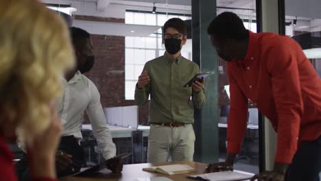 Diverse-group-of-business-colleagues-wearing-masks-brainstorming-in-meeting-room-using-tablet