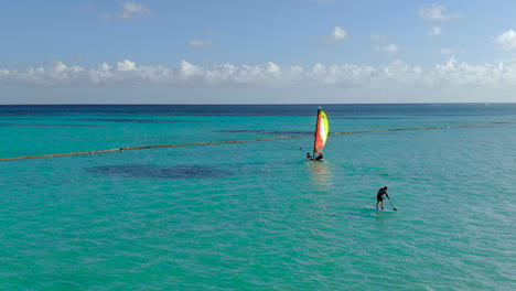 aerial of a paddle border passes behind a small sailboat, dominican republic