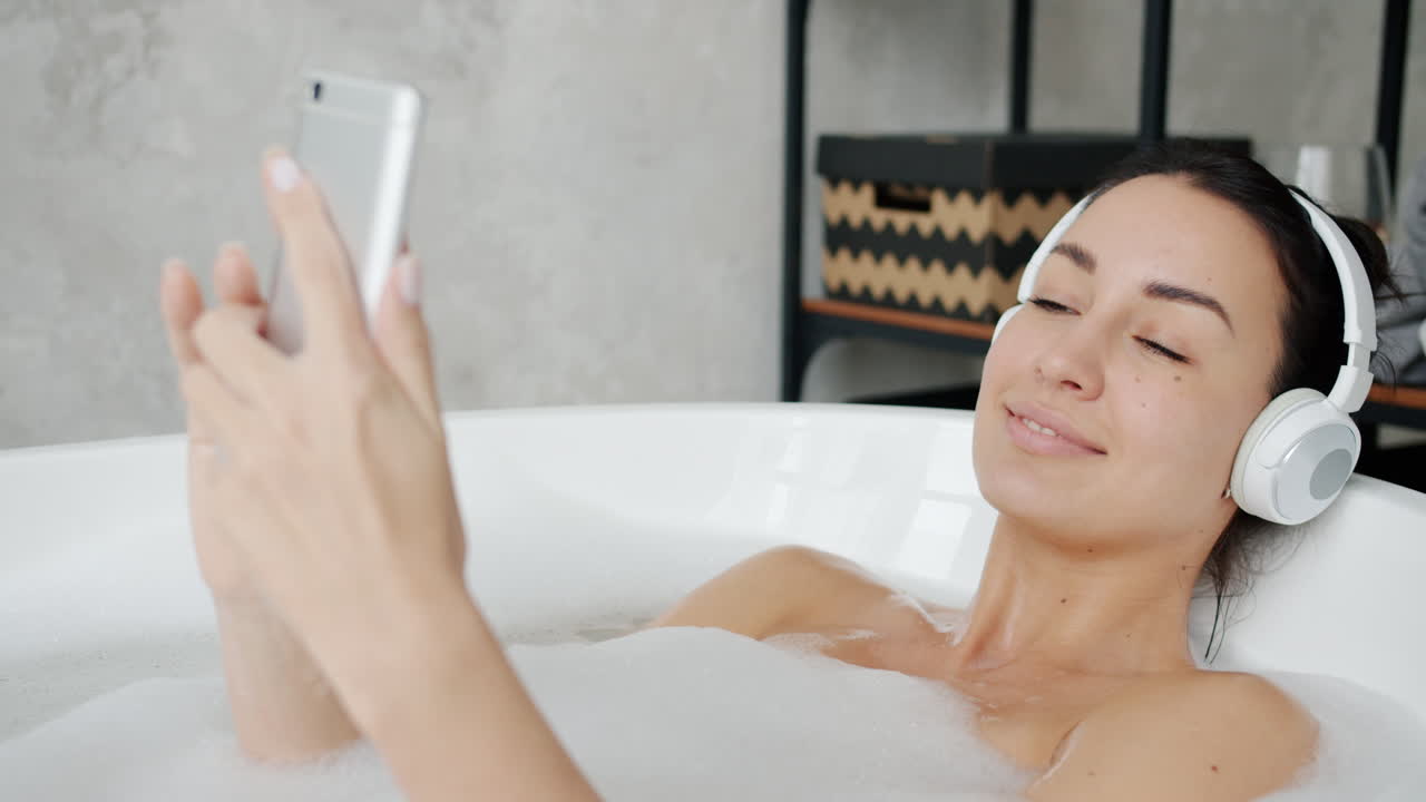 Woman relaxing in a bubble bath with headphones and a smartphone