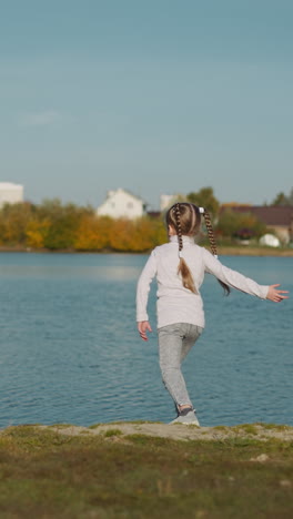 una niña de preescolar con trenzas largas lanza guijarros al río. un niño feliz y activo descansa en la naturaleza. un niño despreocupado se divierte en la orilla verde en un día soleado