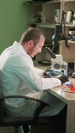 una vista cercana de un técnico en una bata de laboratorio blanca relajándose en su asiento mientras examina cuidadosamente un circuito bajo un microscopio en su laboratorio, con la pared pintada de verde y un estante al lado