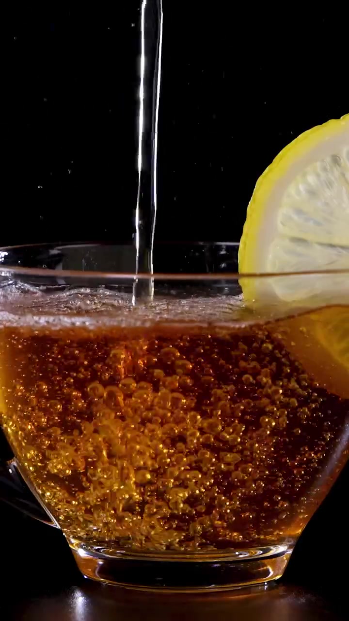 Un getto di tè viene versato in una tazza di vetro con una fetta di limone, creando bolle su uno sfondo scuro.