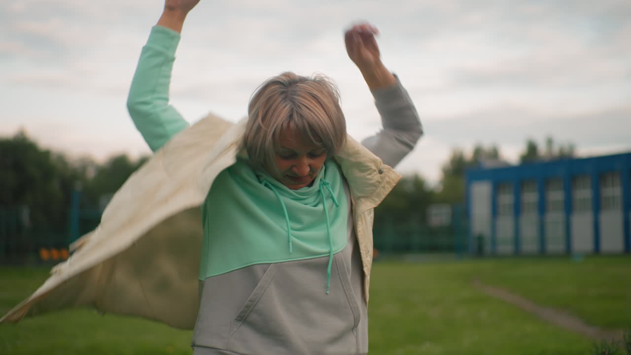 Middle aged woman in mint green hoodie putting on gray jacket while standing outdoors in grassy field near buildings on cloudy day, looking around with relaxed and thoughtful expression