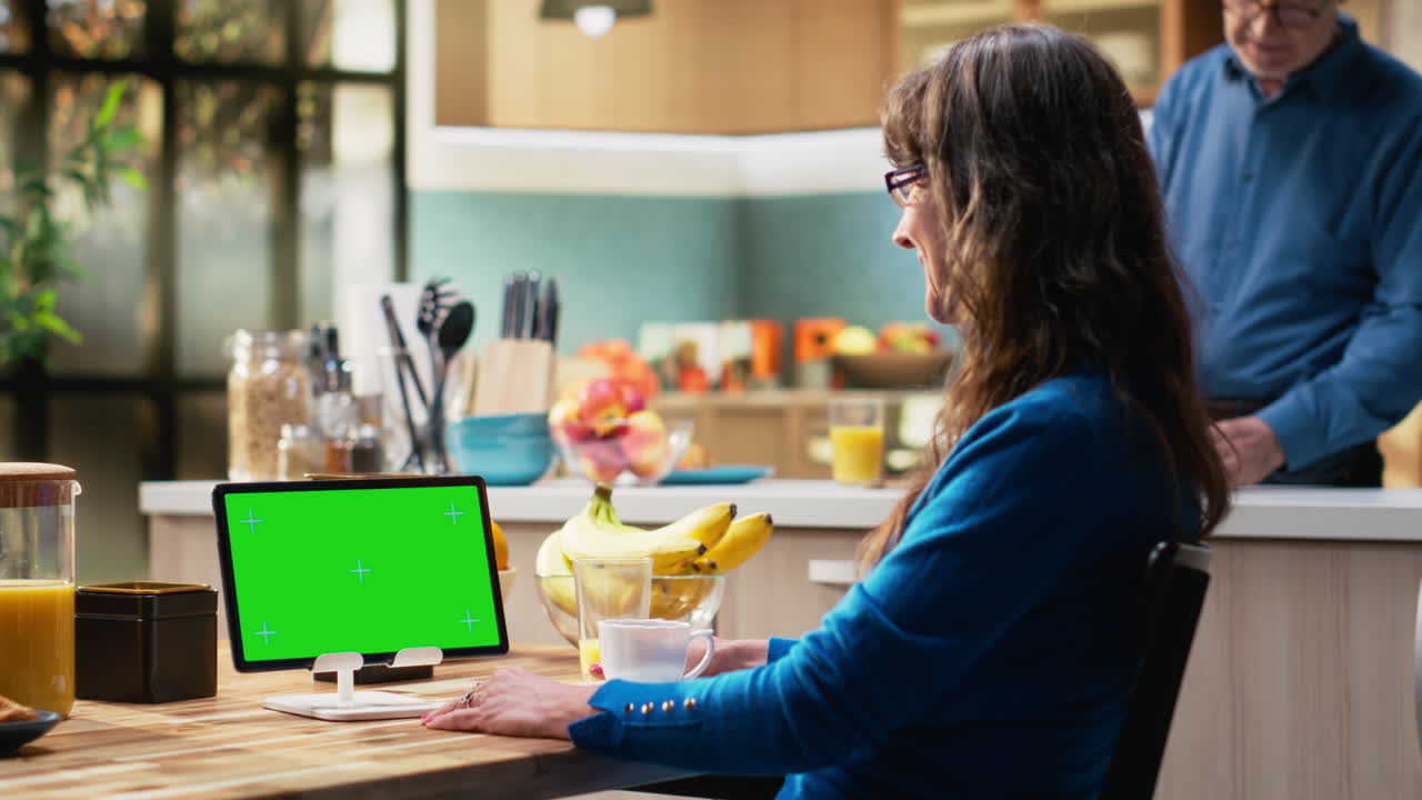 Green screen tablet with senior woman sitting at kitchen table with coffee cups