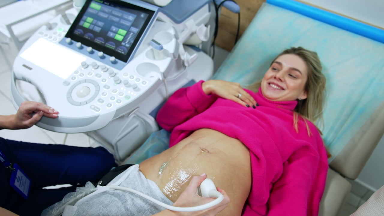 Smiling Caucasian woman in pink sweater is checked on the ultrasonic machine. Woman is happy to see her future child on the monitor. Top view.