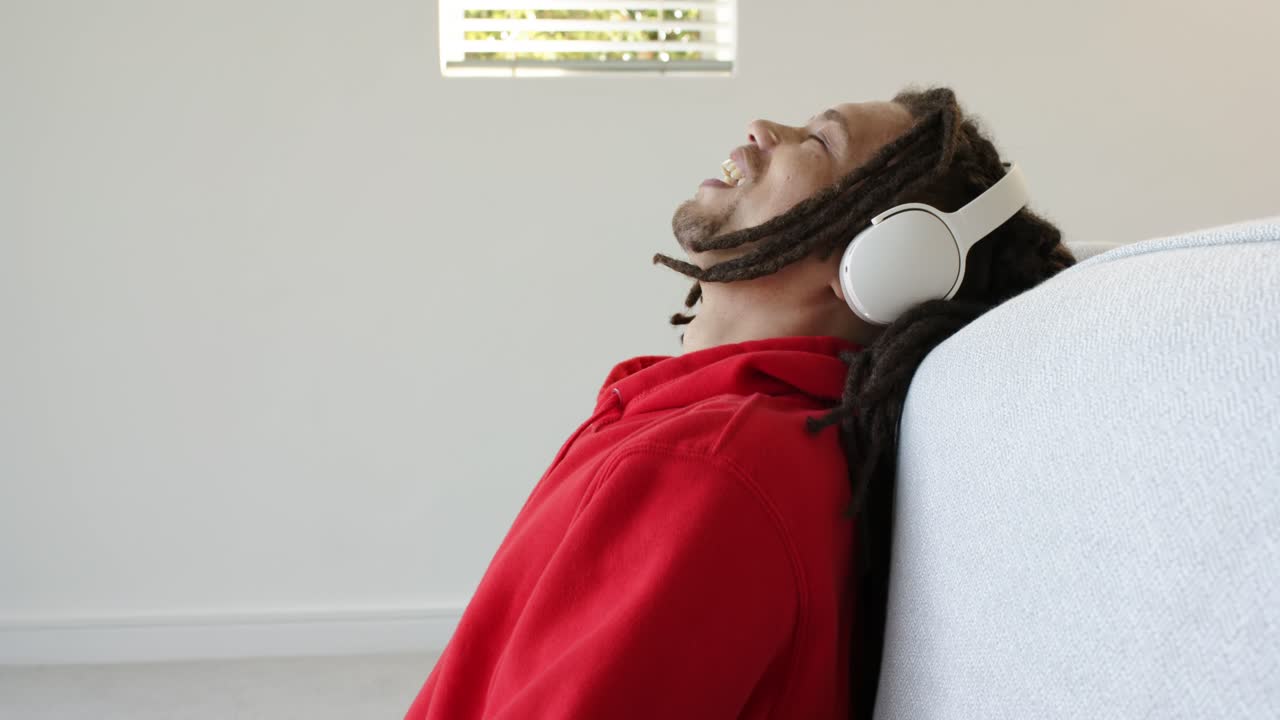 Mid adult African American man in red hoodie headphones scrolling phone resting on sofa relaxing