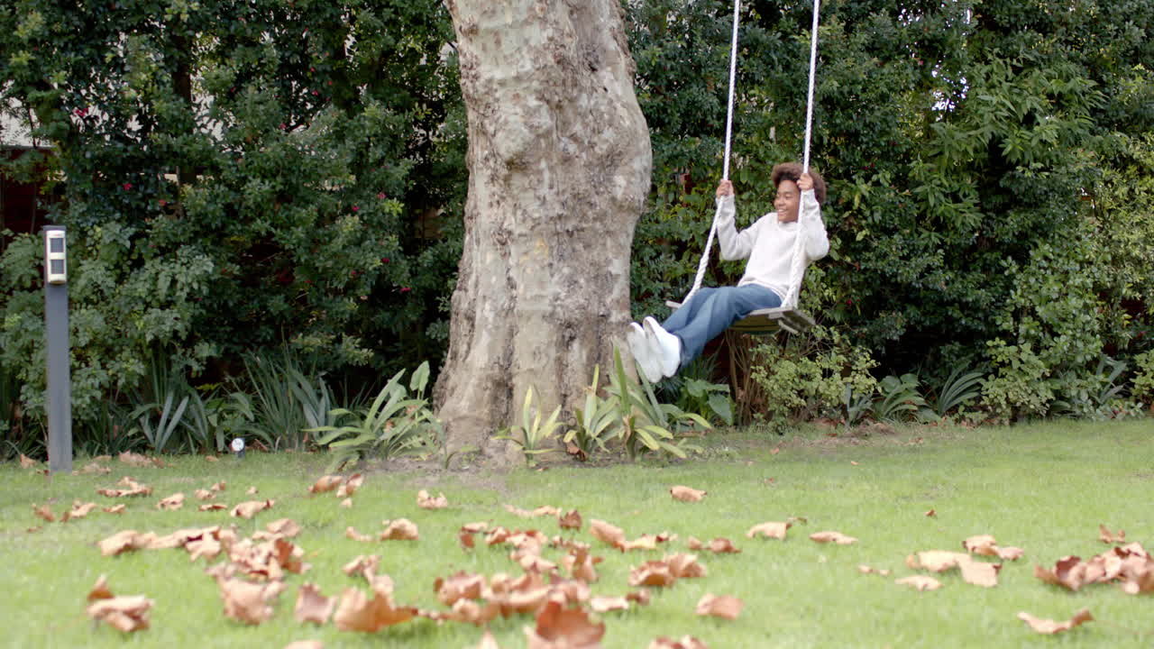 Happy african american boy swinging on tree swing in garden, copy space, slow motion