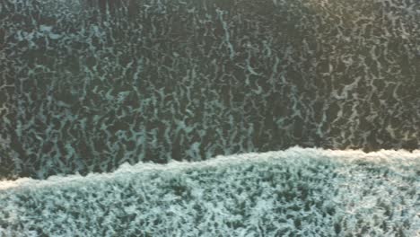 Aerial-tracking-shot-from-left-to-right,-over-waves-crashing-onto-a-sandy-beach