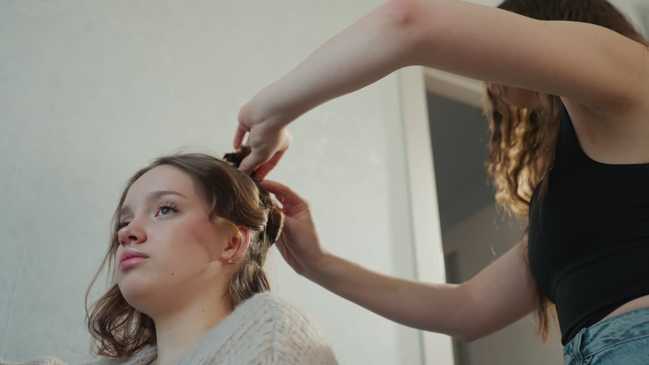 Girl Receives Braid, Friend Gently Braids Girl At Home, Caucasian Woman Relaxing As Companion Adds Braid, Casual Indoor Setting Where Girl Is Being Braided By Friend Amid Soft Natural Light