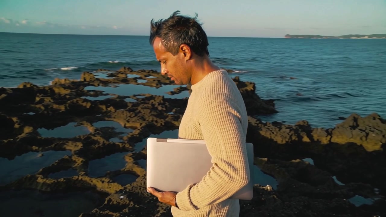 Uomo con maglione beige che sposta il peso camminando tra pozze di marea rocciose, tenendo un laptop argentato per proteggerlo.