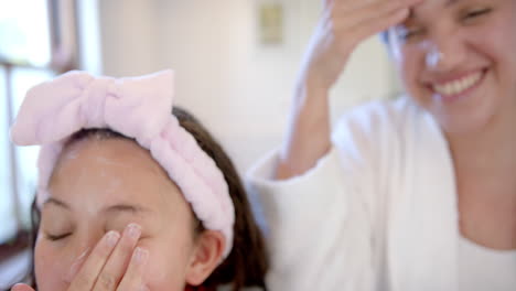 Happy-biracial-mother-and-daughter-with-headbands-putting-cream-on-faces-in-sunny-bathroom