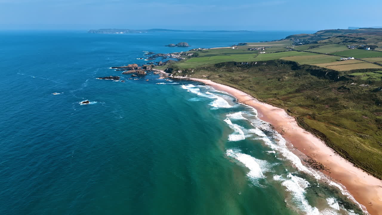 Flying over azure waters of the Pacific Ocean at the coast of Ireland. White waves arrive to the narrow sandy beach. Aerial view.