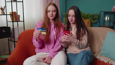 cheerful girls friends using credit bank card and smartphone while transferring money, purchases